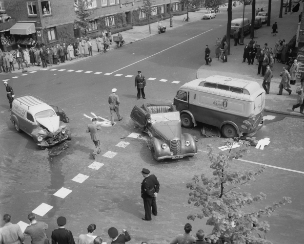 Schwarzes und weißes Bild eines Autounfalls an einer Straßenkreuzung mit mehreren Fahrzeugen, darunter ein Van, und einer Gruppe von Menschen in der Nähe, mit Gebäuden, Bäumen, Pfählen und Texttafeln im Hintergrund.