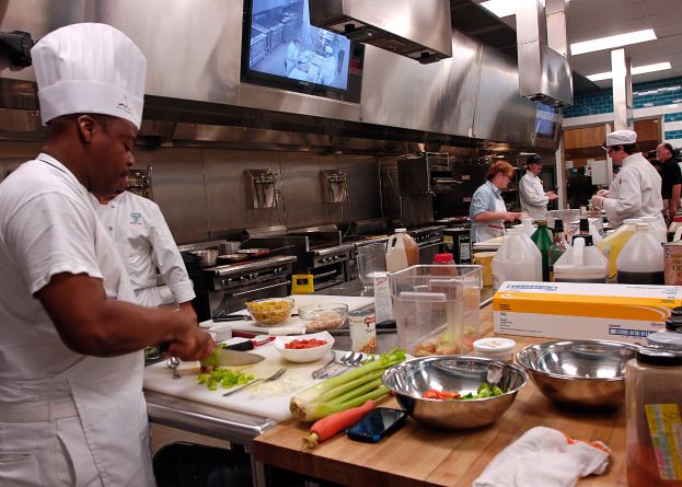 Eine Gruppe von Köchen in einer Küche bei der Zubereitung von Essen auf einem Schneidbrett, mit einem Koch im Vordergrund in einer weißen Schürze und Mütze, der ein Messer hält, umgeben von verschiedenen Utensilien, Schüsseln, Gemüse, einem Mobiltelefon, Flaschen und anderen Gegenständen, mit einem Fernseher und Deckenleuchten im Hintergrund.
