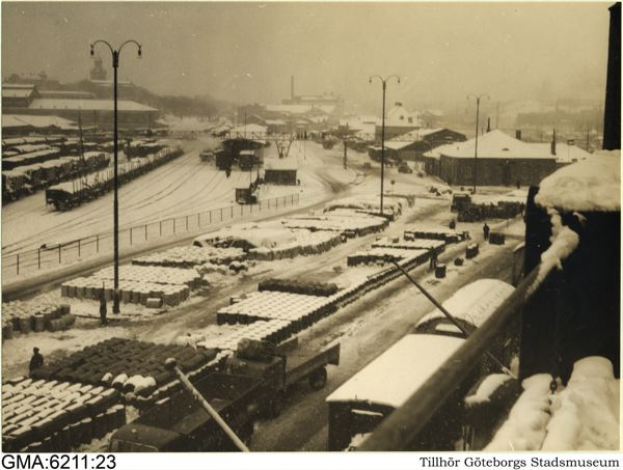Ein Schwarz-Weiß-Foto einer verschneiten Straße mit Fahrzeugen, Masten, Lichtern, Gebäuden, Bäumen und einem Zaun, mit Text am unteren Rand.
