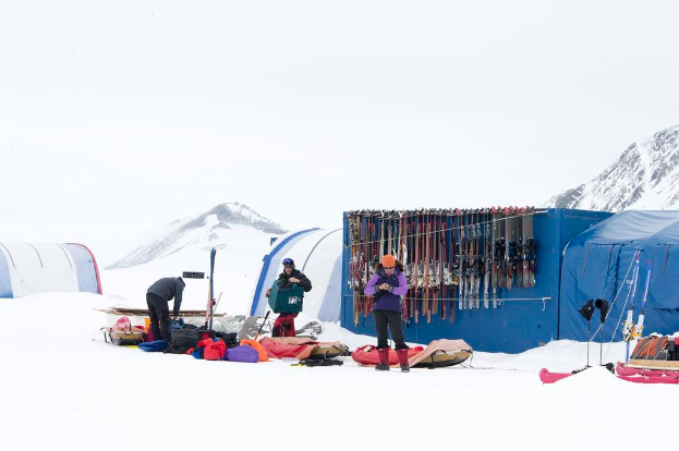 Drei Personen stehen auf einer verschneiten Landschaft mit Taschen herum, Zelte mit Skiern dahinter und schneebedeckte Hügel im Hintergrund unter einem klaren Himmel.