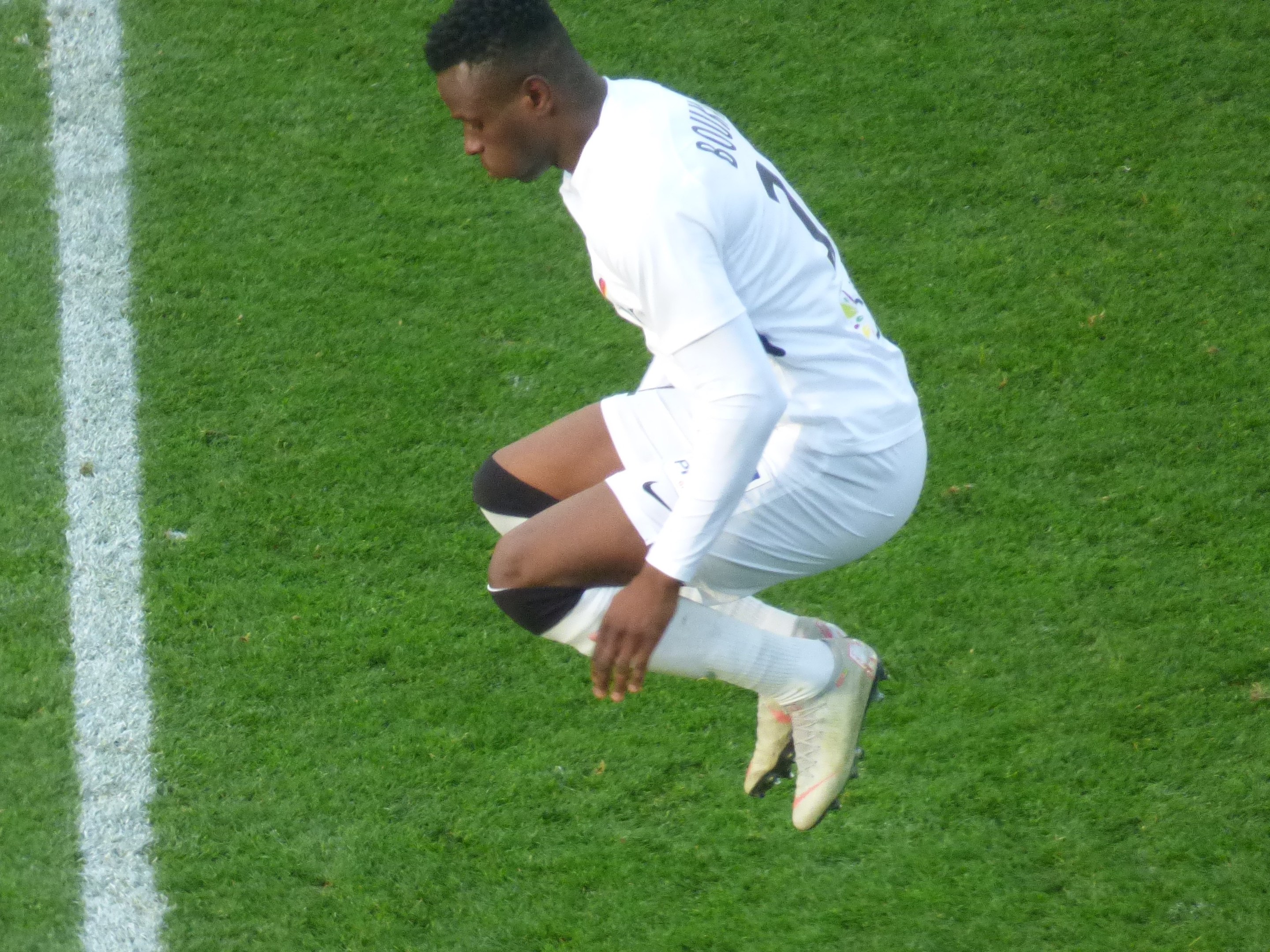 Ein professioneller Fußballspieler in weißer Uniform kniet auf einem Fußballfeld, trägt Fußballstiefel.