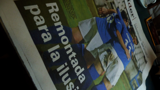 Eine Zeitung und ein Unterteller auf einem Tisch, wobei die Zeitung drei Männer in blauen Hemden zeigt, die möglicherweise Fussball spielen, und Text am unteren Rand des Bildes.