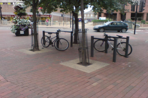 Eine Szene mit Fahrrädern, Bäumen, einem Zaun, einem Auto und Gebäuden.