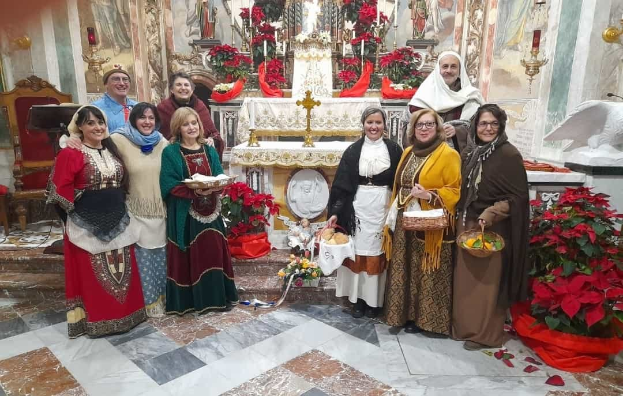 Eine Gruppe von Menschen steht vor einer Kirche und hält Körbe und Teller in den Händen, mit Blumenarrangements, Statuen, Kerzenständern und Gemälden im Hintergrund während einer Weihnachtsfeier.