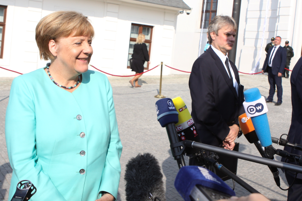 Bundeskanzlerin Angela Merkel spricht vor einem Gebäude mit Fenstern zu Reportern, umgeben von einer Gruppe von Menschen, einige halten Mikrofone auf Stativen, mit einem Seil, das an einen Pfahl gebunden ist, und einem bewölkten Himmel.