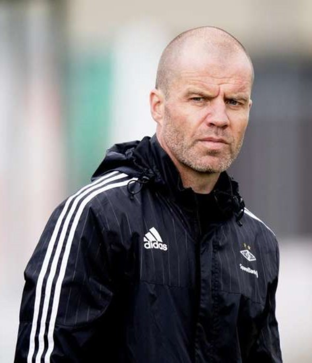 Ein kahlköpfiger Mann in einer blauen Jacke mit weißen Streifen und einem weißen Kragen steht auf einem Fußballfeld, die Hände verschränkt, mit einem ernsten Gesichtsausdruck.