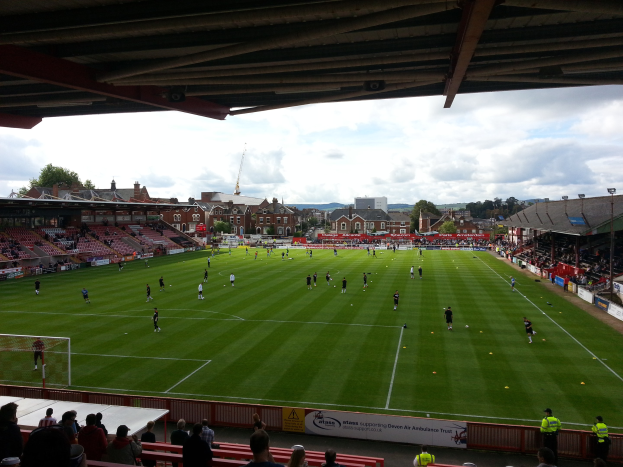 Ein Fußballspiel im St. Mary's Stadium mit Zuschauern auf den Tribünen und auf dem Rasen, einem Torpfosten auf der linken Seite und im Hintergrund Gebäuden, Bäumen und einem bewölkten Himmel.