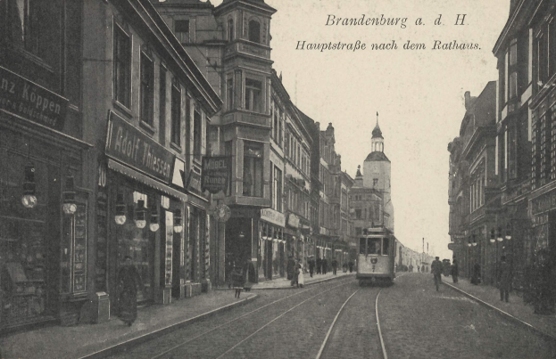 Schwarz-weiß-Foto einer Straßenkreuzung in Brandenburg, Deutschland mit einer Tram auf den Schienen, Fußgängern, Gebäuden mit Fenstern, Schildern, Straßenlaternen und einem bewölkten Himmel.