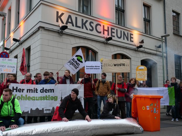 Eine Gruppe von Menschen demonstriert vor einem Gebäude in Deutschland, mit Schildern und Plakaten, wobei zwei Personen im Vordergrund sitzen und ein Müllcontainer rechts zu sehen ist.