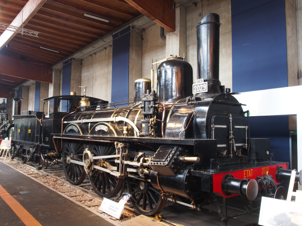 Alte Dampflokomotive in einem Museums Setting umgeben von Eisenbahnschienen, Informationsschildern, Ständen und anderen Ausstellungsstücken, mit einer Wand und Deckenbeleuchtung im Hintergrund.