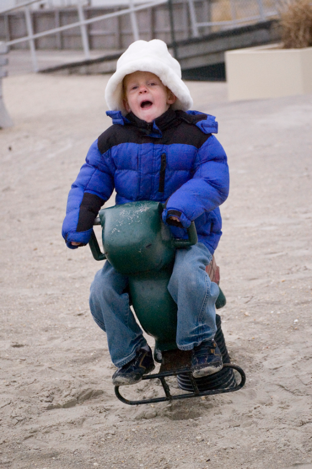 Ein kleiner Junge in einer blauen Daunenjacke sitzt auf einem eisernen Federsessel mit einem weißen Rohrgeländer im Hintergrund.