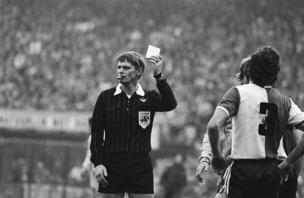 Schwarzes und weiße Bild eines Schiedsrichters mit einer roten Karte vor einer Menge von Fürballspielern, mit einem Zaun im Hintergrund.