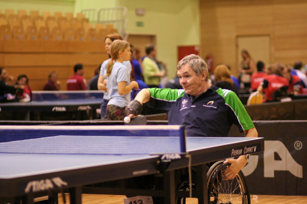 Ein Mann im Rollstuhl spielt Tischtennis, hält den Rollstuhl mit einer Hand und einen Schläger mit der anderen, mit anderen Spielern und Tischen im Hintergrund.