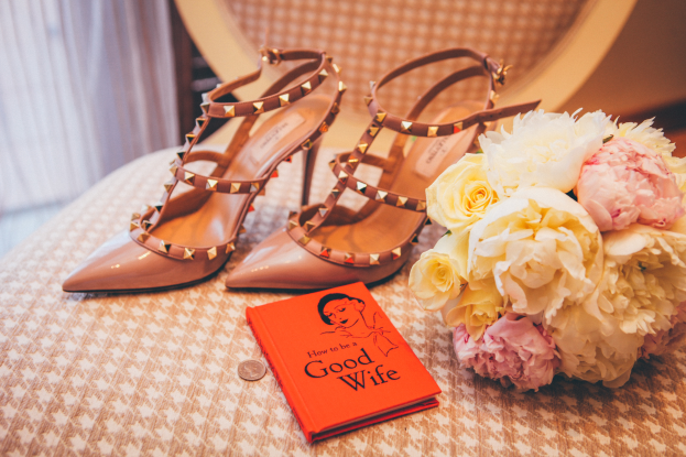 Ein Tisch zeigt ein Buch mit dem Titel "Wie man eine gute Ehefrau ist", einen Blumenstrauß und ein Paar Valentino-Schuhe, vor einem Vorhang im Hintergrund.
