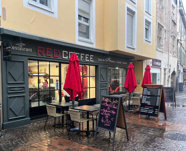 Ein rotes Kaffeehaus an einer Straßenecke mit Außensitzplätzen unter roten Schirmen, schwarze Schilder mit Text, Gebäude, Bäume und einen klaren blauen Himmel im Hintergrund.