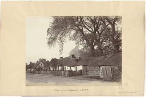 Schwarzes und weißes Foto eines Dorfs mit Reetdächern, einer Straße, einem Zaun, Bäumen und einem Himmel im Hintergrund, mit Text unten.