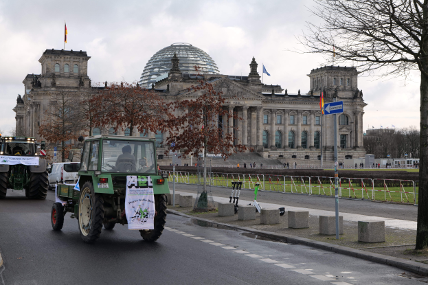 Eine Gruppe von Traktoren f√§hrt die Stra√Ÿe vor dem Reichstaggeb√§ude in Berlin, Deutschland, entlang, das mit Fenstern, S√§ulen und Flaggen geschm√ľckt ist und von B√§umen, Gel√§ndern, Pfosten und anderen Gegenst√§nden umgeben ist.