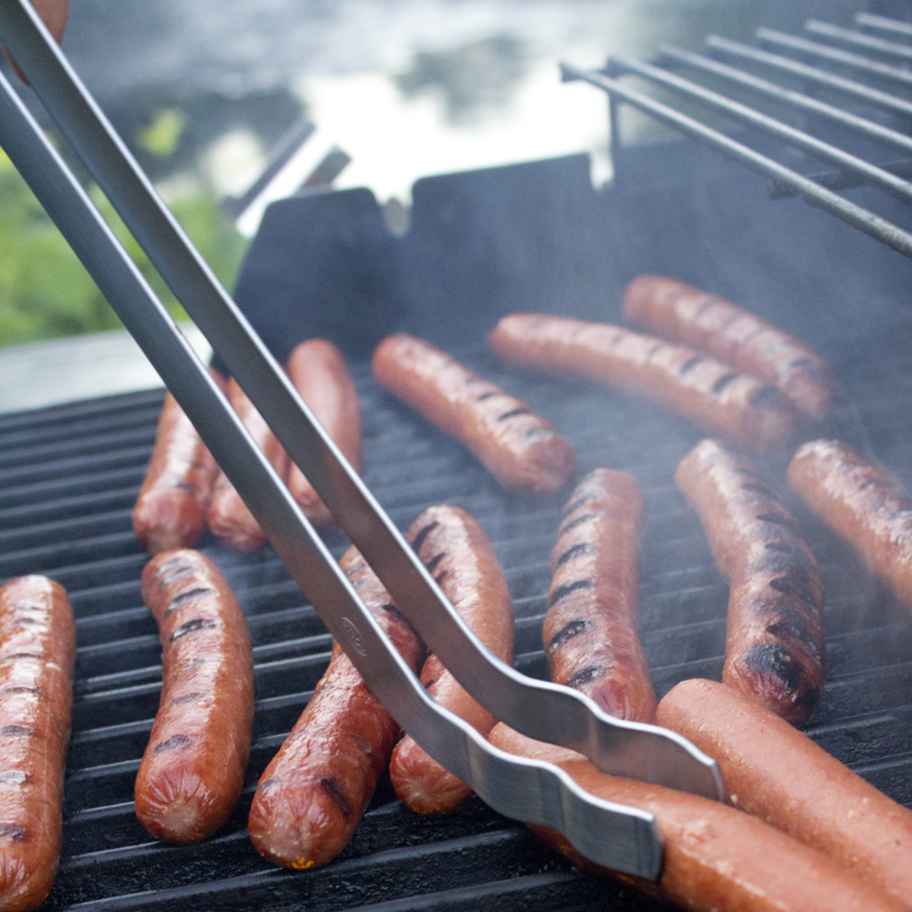 Würstchen auf einem Grill mit Zange, eine Person Hand und Pflanzen im Hintergrund bei Tag.