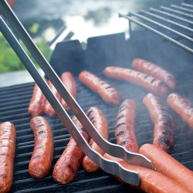 Würstchen auf einem Grill mit Zange, eine Person Hand und Pflanzen im Hintergrund bei Tag.