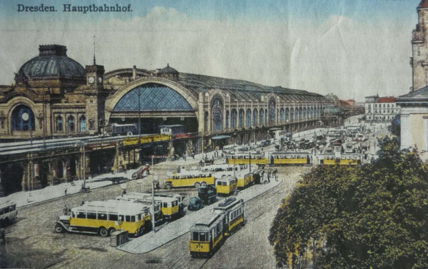 Eine alte Postkarte zeigt einen belebten Dresdner Bahnhof mit Fahrzeugen, Menschen, Bäumen, Gebäuden und einem bewölkten Himmel, mit der Aufschrift "Dresden Hauptbahnhof" oben drauf.