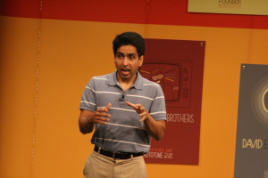 Ein Mann steht auf einer Bühne und spricht vor einer Wand mit Plakaten.