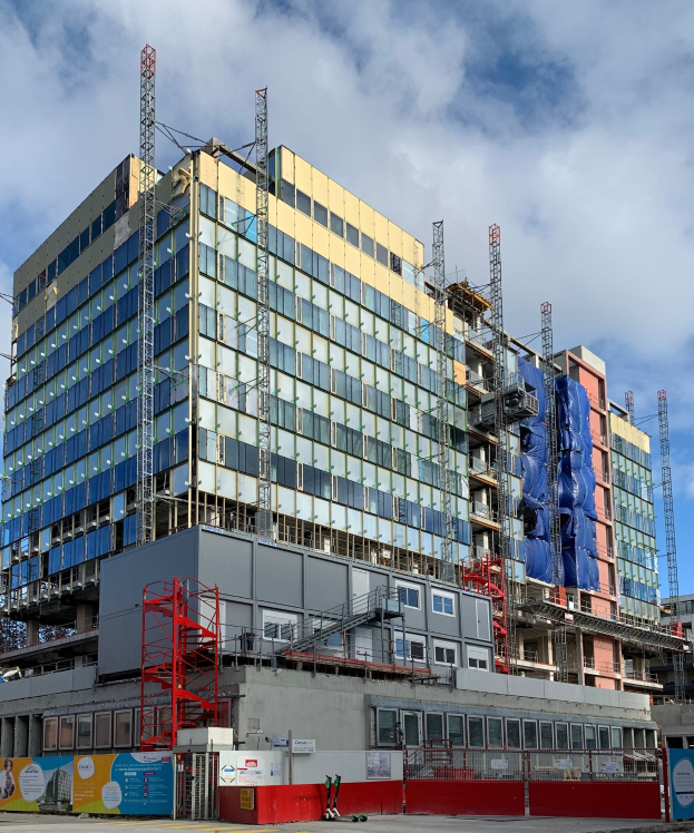 Ein großes städtisches Gebäude im Bau mit Glasfenstern, Metallrahmen, einer Treppe, einem Zaun, einem Schild, Pfosten, einem Straßenpfosten, umgeben von Bäumen und einem bewölkten Himmel.