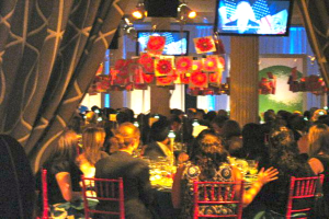 Eine Gruppe von Menschen an Tischen mit Essen, Fernsehern, Lampen, hängenden Papieren und Vorhängen in einem Raum.