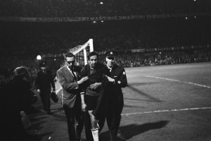 Eine Gruppe von Männern, die auf einem Fußballfeld mit einem Torpfosten und beleuchteten Rängen feiern, in Schwarz-Weiß aufgenommen.