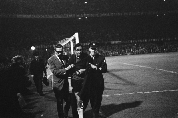 Eine Gruppe von Männern, die auf einem Fußballfeld mit einem Torpfosten und beleuchteten Rängen feiern, in Schwarz-Weiß aufgenommen.