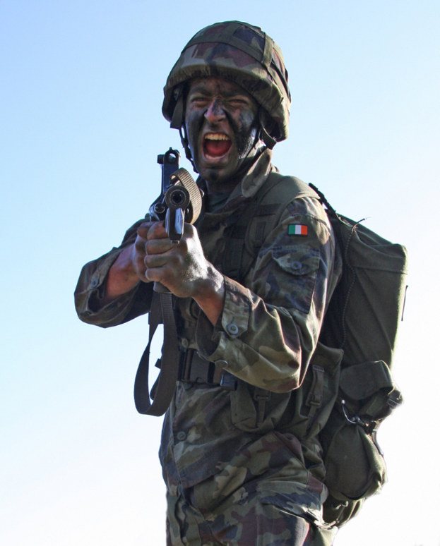 Eine Person in einer Armeeuniform, die ein Abzeichen, Helm und eine Tasche trägt und eine Waffe hält.