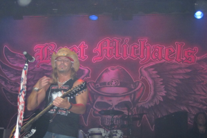 Eine Musikkonzertszene mit einem Gitarristen links neben einem Mikrofon, einer Person in der unteren rechten Ecke, einem Banner im Hintergrund und Drogen in der Mitte unten, alles unter Bühnenbeleuchtung von oben.