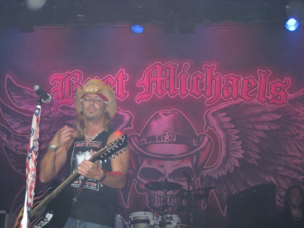 Eine Musikkonzertszene mit einem Gitarristen links neben einem Mikrofon, einer Person in der unteren rechten Ecke, einem Banner im Hintergrund und Drogen in der Mitte unten, alles unter Bühnenbeleuchtung von oben.
