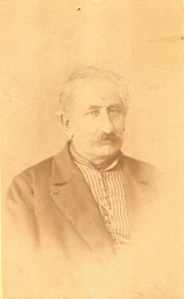 Ein Schwarz-Weiß-Porträt eines älteren Mannes mit Schnurrbart, der einen Mantel trägt und ein starkes Kinn, eine prominente Nase, dunkle Augen und einen nachdenklichen Ausdruck mit leicht geöffneten Lippen zeigt.