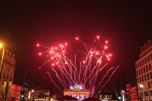 Eine Straßenansicht in Berlin an Silvester, mit Häusern, Bäumen, Laternenmasten, Ampeln, Schildern, Zelten und Menschen, vor einem von Feuerwerk erhellten Himmel.