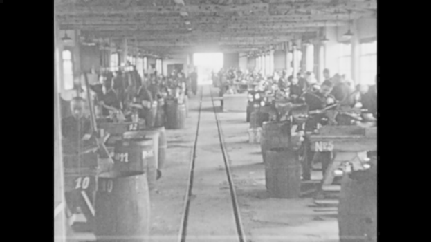 Ein Schwarz-Weiß-Foto einer Fabrik mit Arbeitern an Maschinen, Fässern mit Nummern darauf, einer Eisenbahnschiene, Säulen und Deckenlampen, die an die industrielle Revolution erinnern.