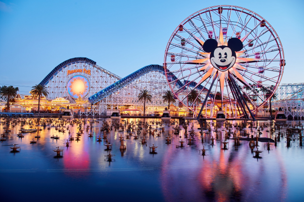 Eine lebendige Dämmerungsszene im Disneyland California Adventure mit einem riesigen Mickey-Maus-Rad im Vordergrund, einer Achterbahn im Hintergrund und Booten auf dem Wasser, mit einem Himmel aus Orangen, Pinks und Purpur und silhouettenartigen Bäumen.