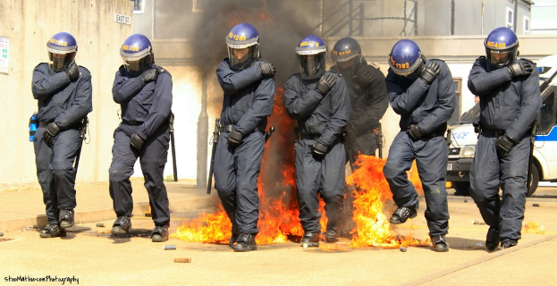 Menschen in Helmen vor einem Feuer, mit Gebäuden und einem Fahrzeug im Hintergrund, einem Plakat und einer Tafel an der linken Wand und Text am unteren Bildrand.