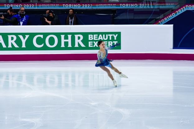 Eine Frau in einem blauen Kleid fährt auf einem Eisstadion während eines professionellen Eiskunstlauf-Events, mit einer Menge von Menschen mit Kameras und einem Schild mit der Aufschrift 'Tessa Virtue und Scott Moir' im Hintergrund.