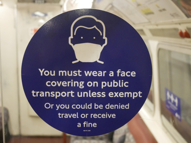 Ein Schild an einer Zugtür informiert Fahrgäste darüber, dass sie im öffentlichen Verkehr eine Gesichtsbedeckung tragen müssen, andernfalls drohen Reiseverweigerung oder Bußgelder, dargestellt in einem Glasfenster.