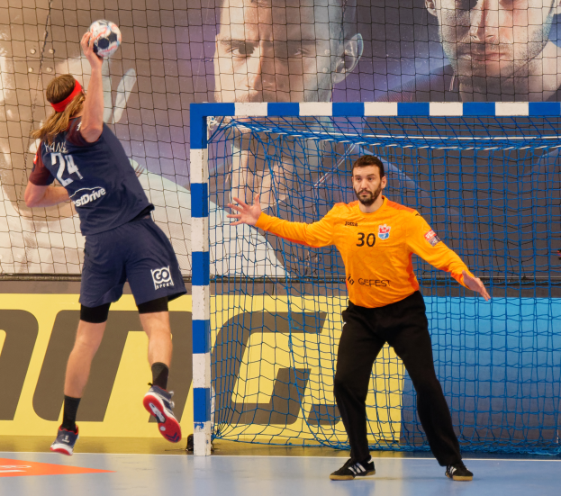 Zwei Männer spielen Handball vor einem Tor mit Netz und Banner im Hintergrund.