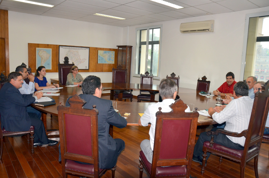 Eine Gruppe von Menschen, die an einem Tisch sitzen und Papiere und Gläser vor sich haben, mit einer Klimaanlage, einer Anschlagtafel und Karten an der Wand im Hintergrund.