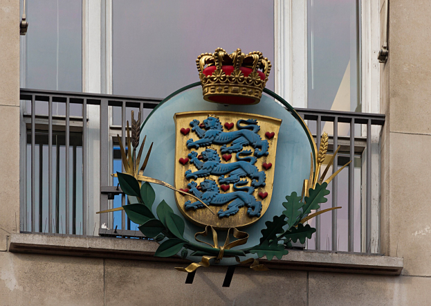 Wappen der britischen Königsfamilie auf einem Gebäudeäußeren mit einer Krone- und Blätter-Logo, Fenstern und Metallgittern im Hintergrund.