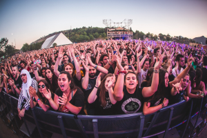 Eine große Menschenmenge auf einem Musikfestival mit erhobenen Händen, die vor einem Geländer stehen, mit Bäumen, Gebäuden und einem klaren blauen Himmel im Hintergrund.