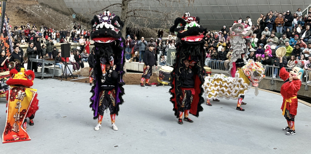 Eine Gruppe von Menschen in Kostümen mit Musikinstrumenten performs auf einer Straße vor einem Publikum während einer Feier zum Chinesischen Neujahr, mit Bäumen und einem Gebäude im Hintergrund.