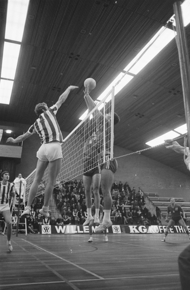 Eine Gruppe von Männern, die Volleyball in einer Turnhalle mit einem Netz, einem Ball in der Luft, Zuschauern auf Treppen und Bannern mit Text im Hintergrund spielen, unter Deckenbeleuchtung.