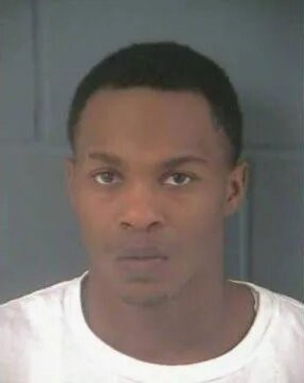 Schwarz-weißes Mugshot eines Mannes in einem weißen T-Shirt, der nach vorne schaut, vor einer neutralen Wand.