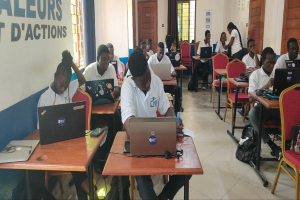 Eine Gruppe von Menschen sitzt an Tischen mit Laptops in einem classroom, mit Taschen auf dem Boden und Text an der linken Wand, was auf einen Online-Kurs in Ghana hinweist.