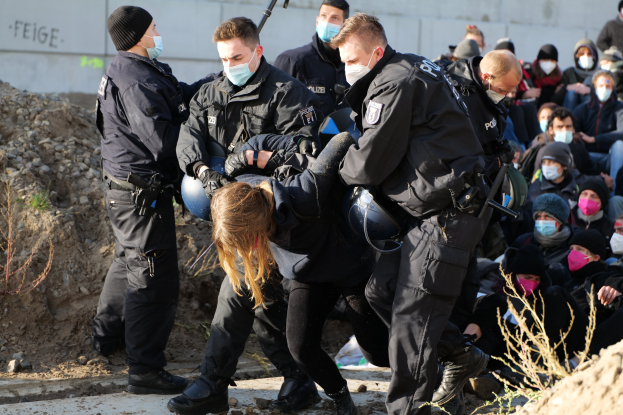 Gruppe maskierter Menschen in Mützen, einige mit Waffen, umgeben eine Frau, die von der Polizei eskortiert wird, mit Pflanzen, Steinen und einer Wand mit Protesttext im Hintergrund.