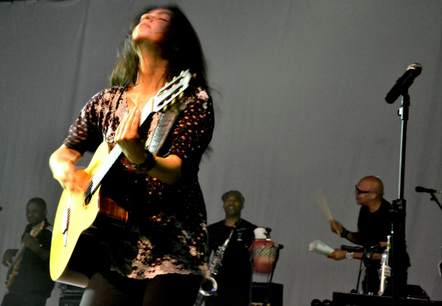 Eine Frau spielt Gitarre auf der Bühne eines Musikkonzerts, umgeben von Musikern und verschiedenen Musikinstrumenten und einem Mikrofon rechts daneben.