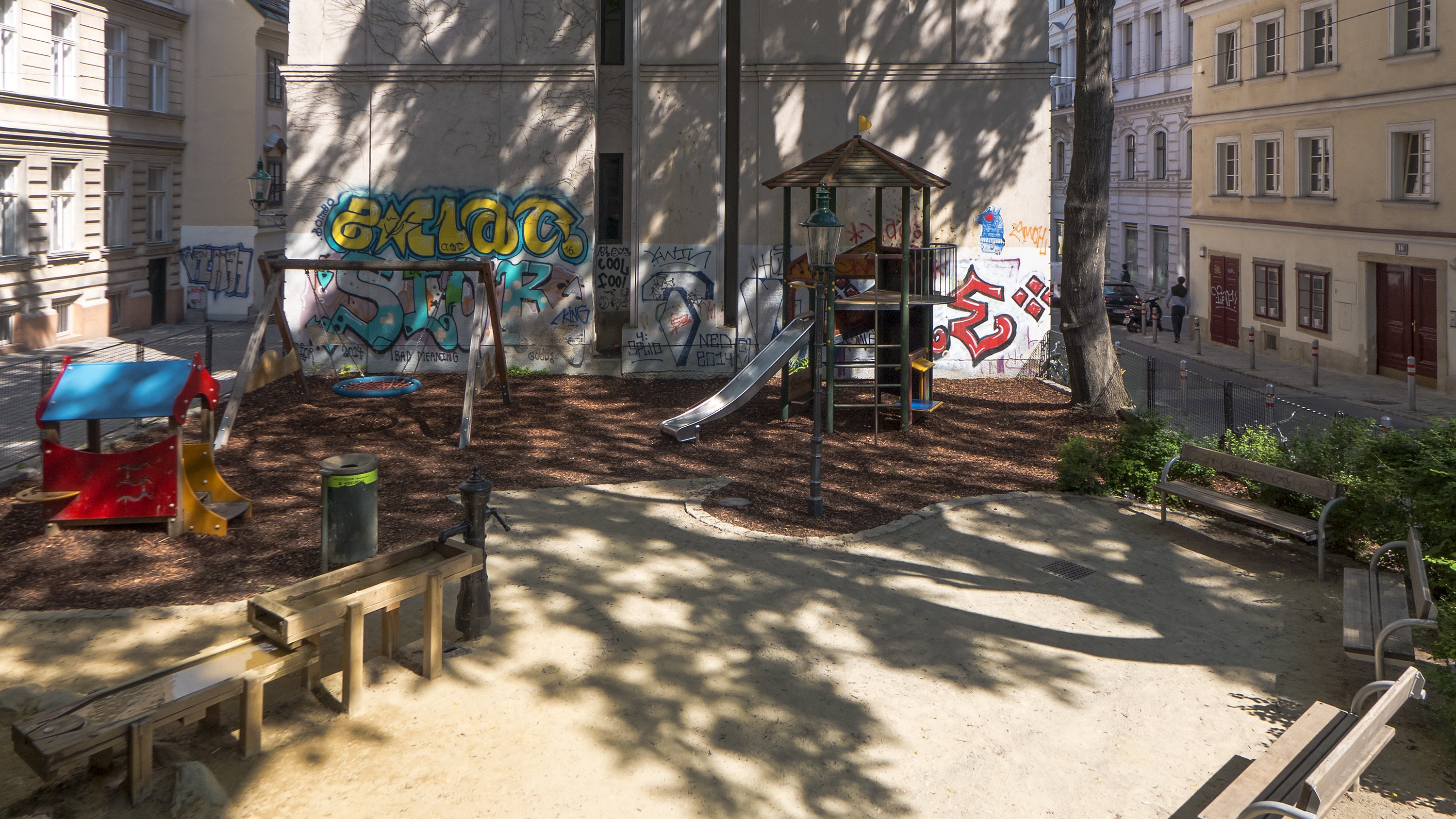 Spielplatz mit Graffiti in einer städtischen Umgebung, umgeben von Gebäuden, Bäumen, Bänken und einer Straße mit Menschen und Fahrzeugen.
